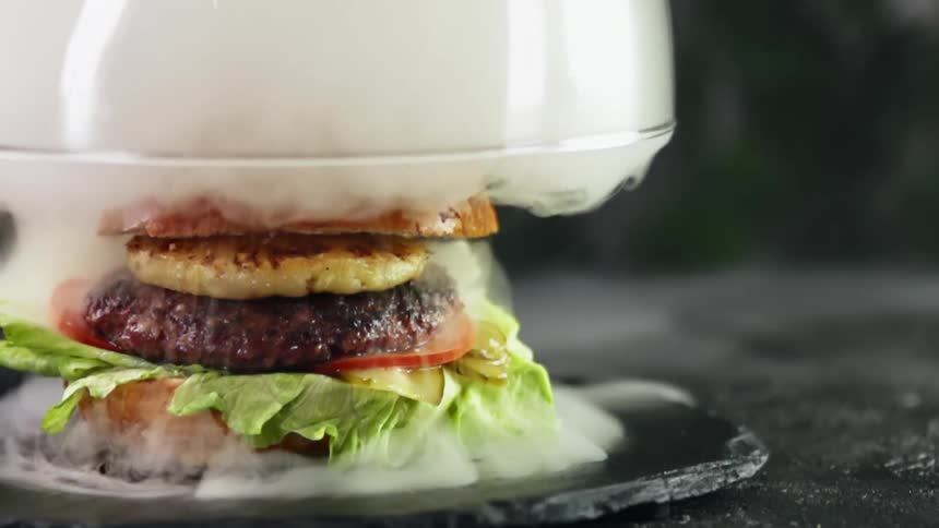 A glass dome filled with smoke is lifted to reveal a gourmet burger with a slice of grilled pineapple