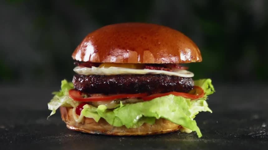 The top of a brioche bun is dropped onto a burger with a fried egg and bacon