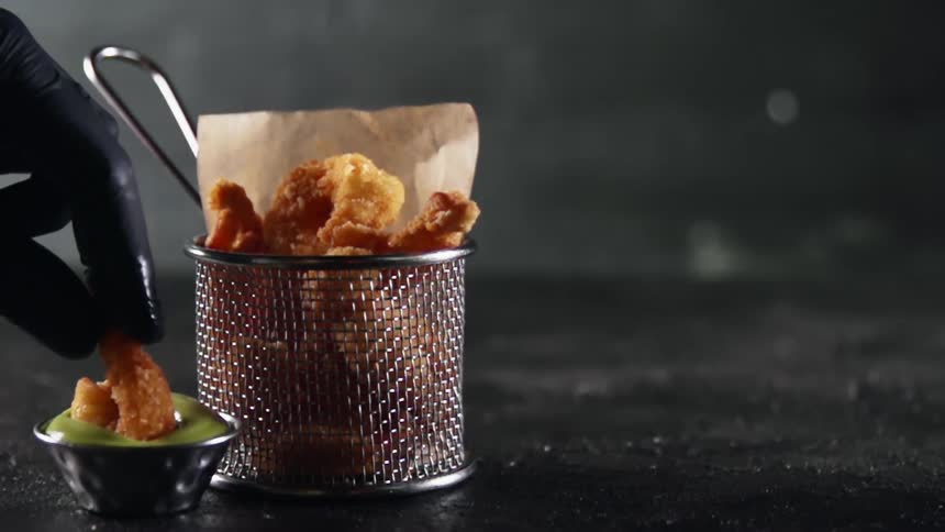 A blacked gloved hand dips a shrimp covered in breadcrumbs from a basket into a green sauce