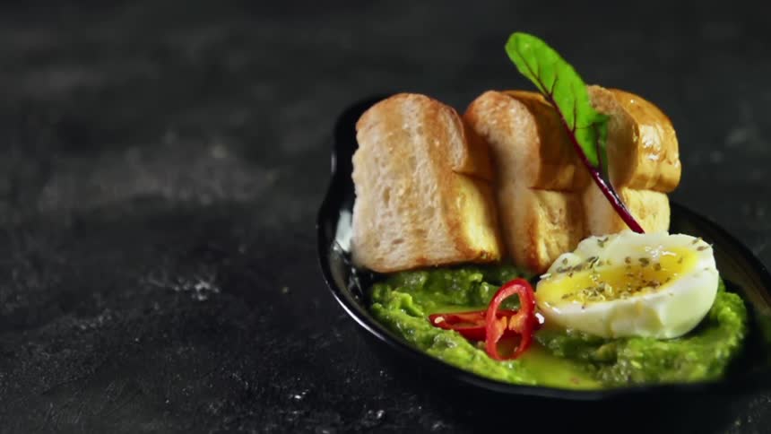 Leafy greens and tomato slices fall on a black ceramic dish containing sliced toasted bread, guacamole and a soft boiled egg