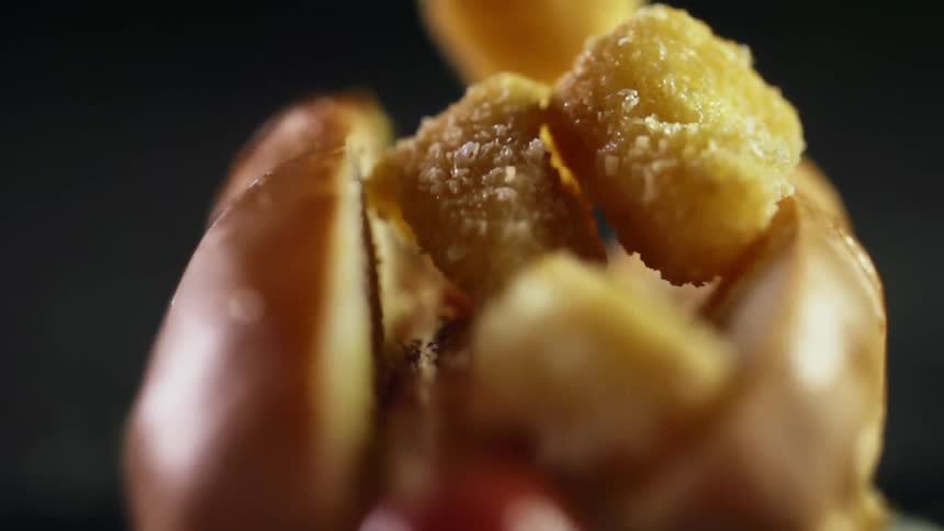 Tiny fried nuggets falling onto a sausage between a hot dog bun