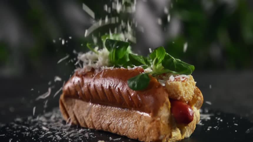 Shredded cheese is dropped onto hot dog topped with greenery