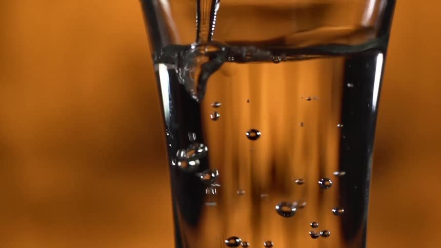 Clear liquor pouring in a short shot glass against a yellow background