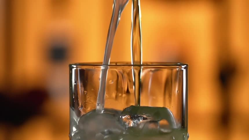 Liquor pouring into a glass filled with ice cubes against a yellow background