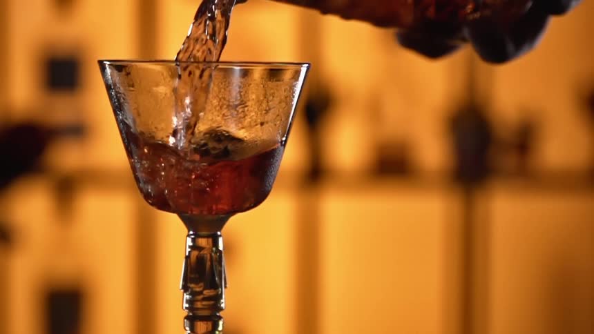 A person is filling up a glass goblet with brownish liquor