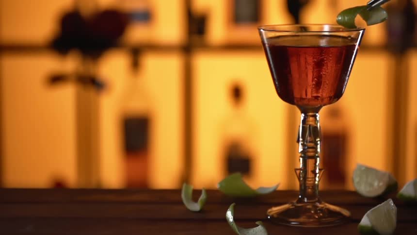 A lime peel being placed on the rim of a drinking glass filled with an amber liquor with a pair of tweezers