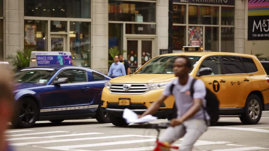 Vehicles driving down a city street with people walking and biking