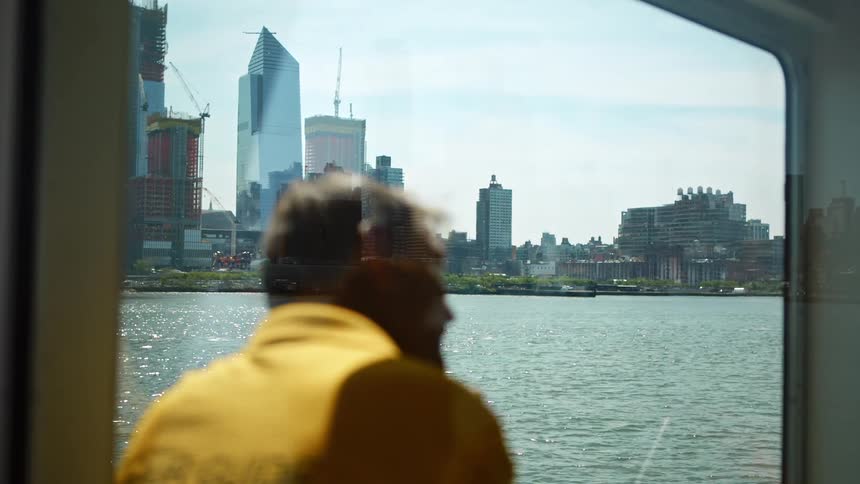 A man sits near a window on a boat in the ocean and looks as the city skyline while chatting