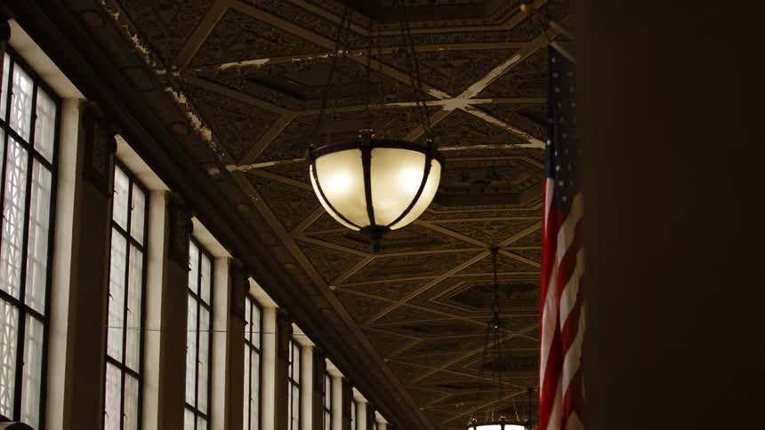 A large american flag hangs inside a building lined with windows along a wall