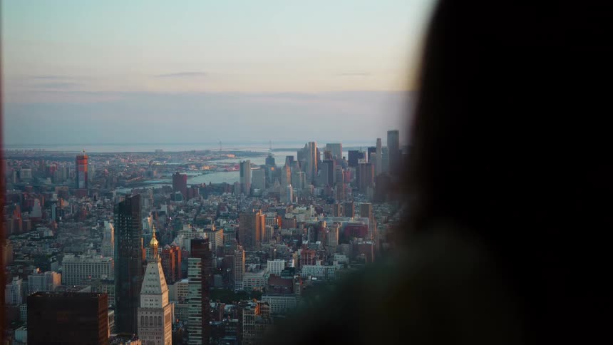Woman looks out the window at big city skyline from a distant with tall buildings and skyscrapers on the horizon at sunset