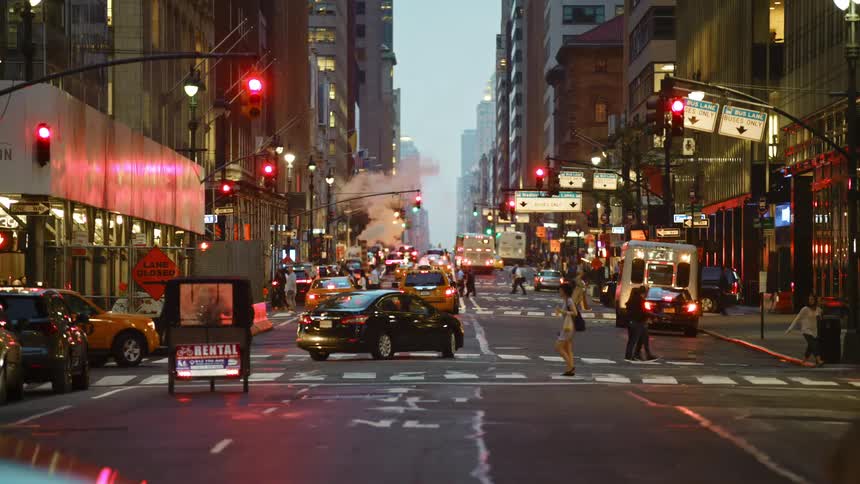 Busy city street with pedestrians crossing a road and vehicles drive on a highway at dusk in downtown