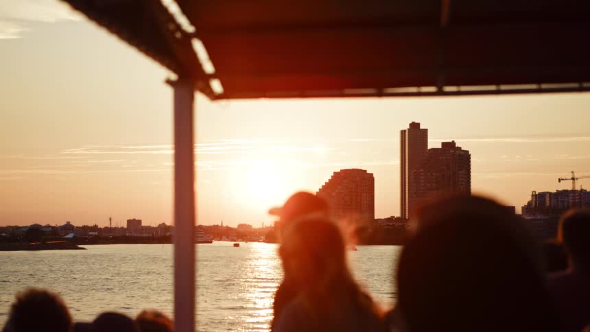 A group of people walk around on a ferry as it moves across a bay near a large city