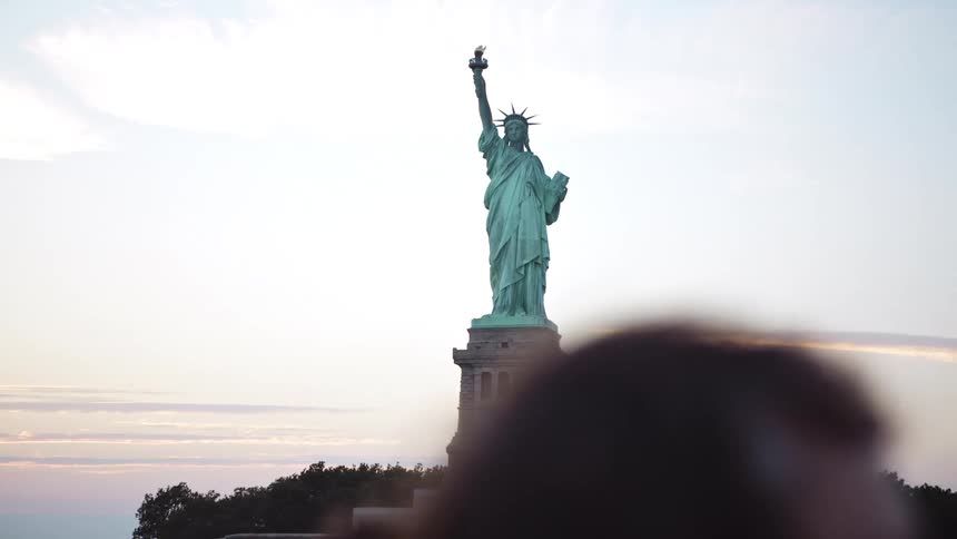 In the distance behind a few tourists the statue of liberty stands tall before a cloudy sky
