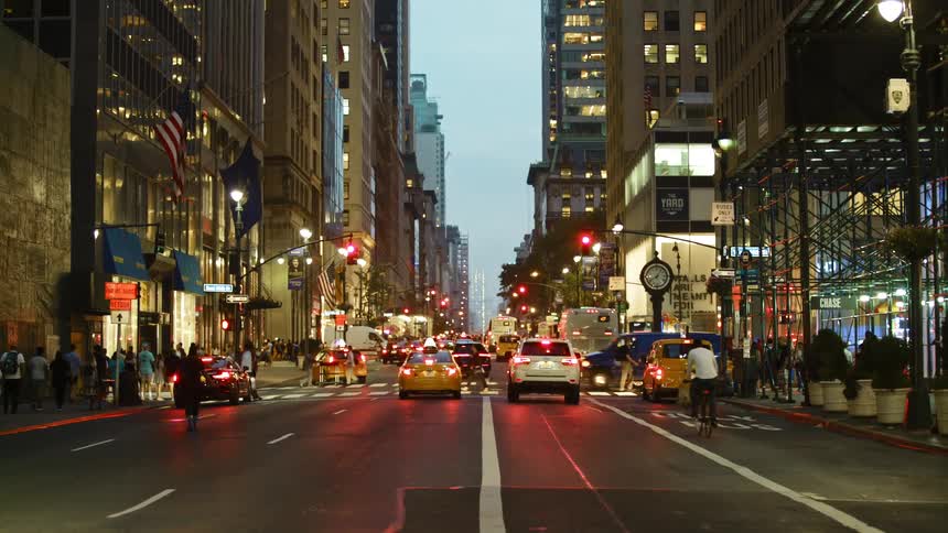 Vehicles sit at a city traffic light with pedestrians surrounding the area