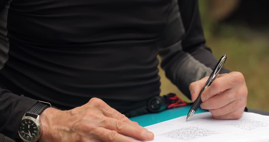 A man sketches using a pen and pad while sitting outside on a clear sunny day