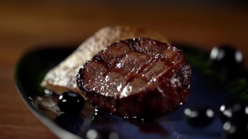 Black olives falling on a plate next to a cooked piece of cooked steak