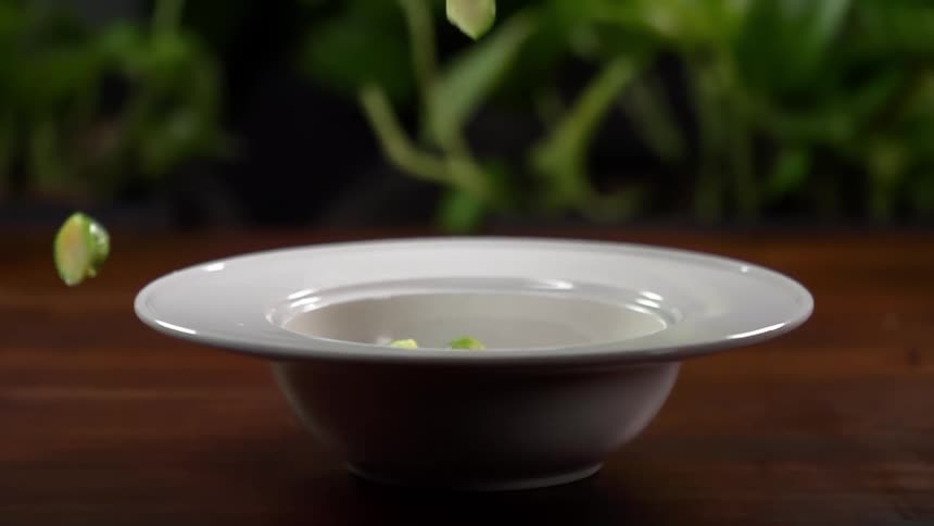Chopped vegetables are dropped into a white bowl and some bounce out onto the table