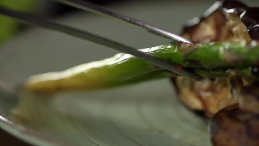 A piece of grilled asparagus is carefully placed on a plate next to grilled zucchini