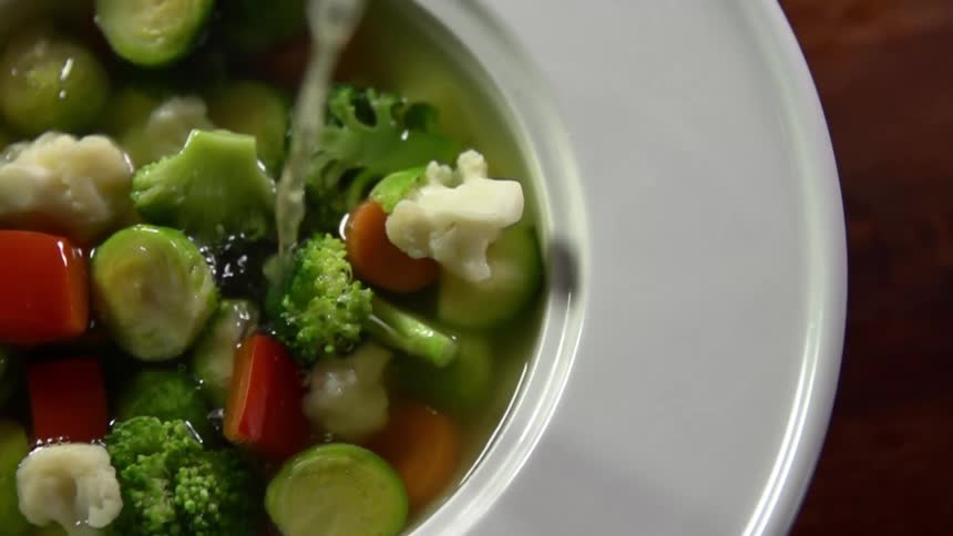 Liquid broth is poured over vegetables in a glass bowl to make a soup