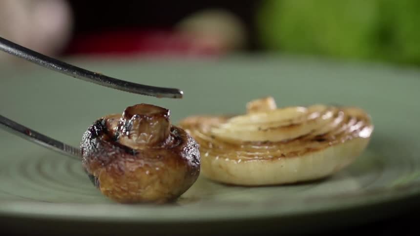 A pair of metal tongs drops a charred mushroom on a plate containing part of an onion