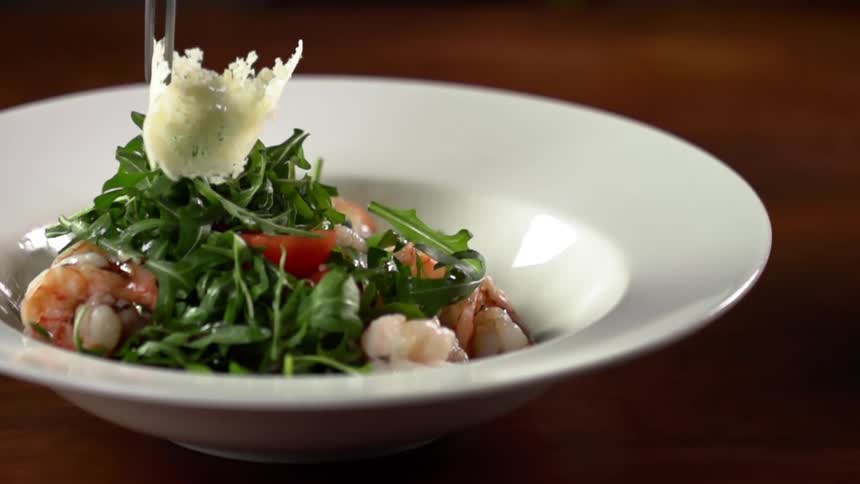 Tongs add tube shaped food to mound of green leafy vegetables topping shrimp in shallow white bowl