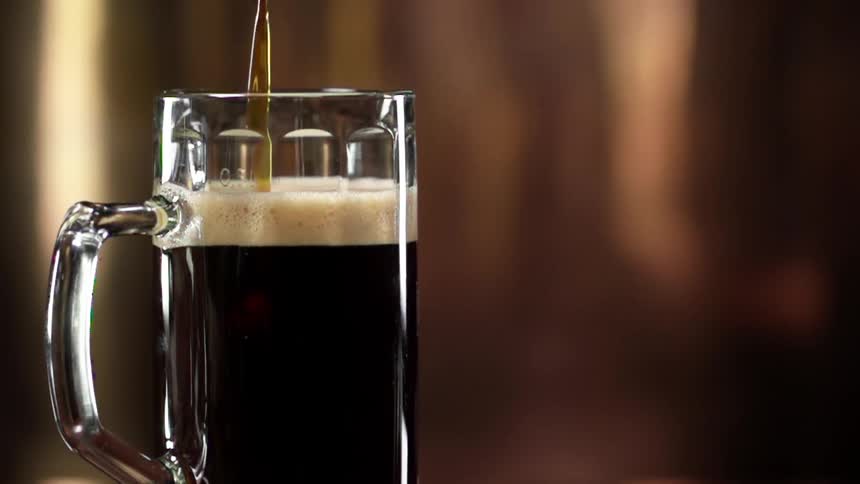 A glass mug is filled with a dark brown ale with a creamy layer of foam atop its surface
