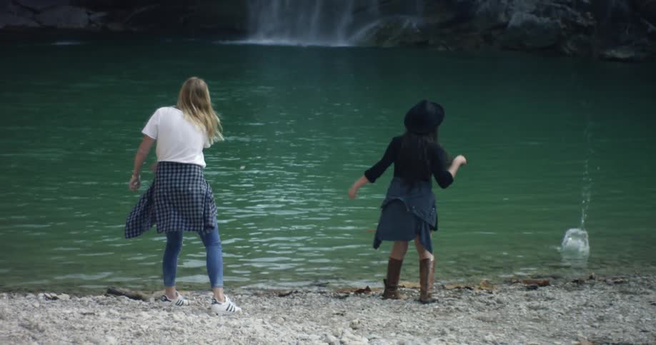 Two women are throwing rocks in the water before a waterfall