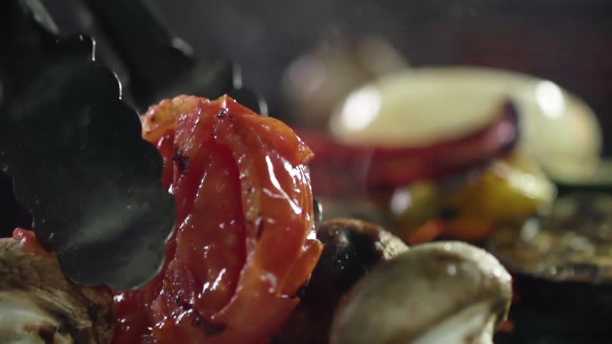 Grilled steaming tomato in front of the fire is picked up by a metalic tong