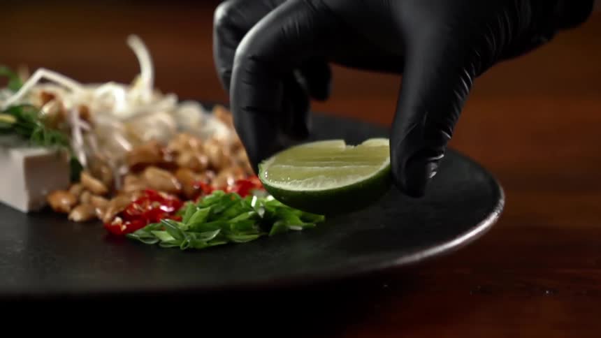 A gloved hand places half of a lime on a black plate next to fresh garnishes and toppings