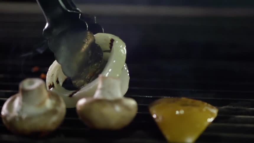 Tongs pick up onion pieces from the grill while mushrooms and peppers continue to cook