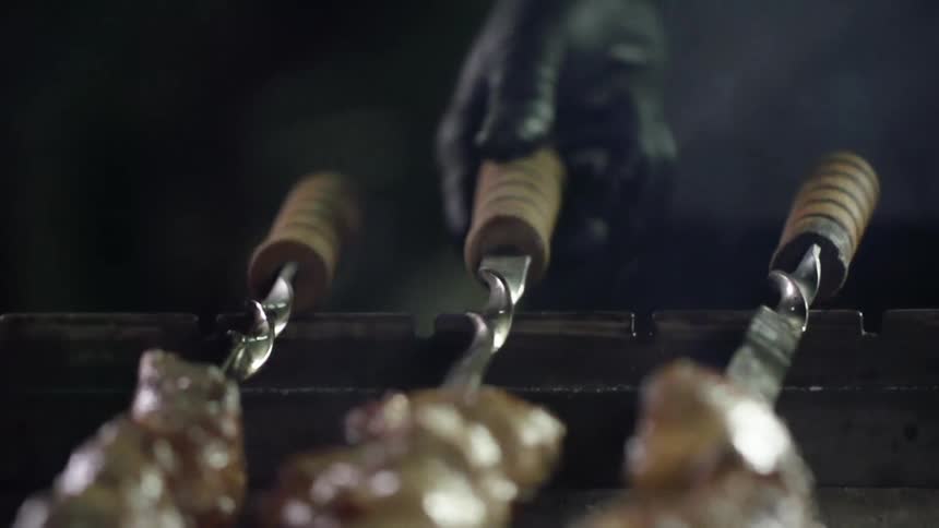 A cook wearing a black glove turns skewers of meat over a grill flame