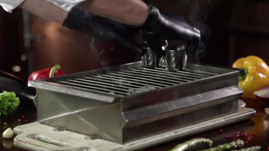A person wearing black disposable gloves slowly places two metal cups onto a small smoking grill