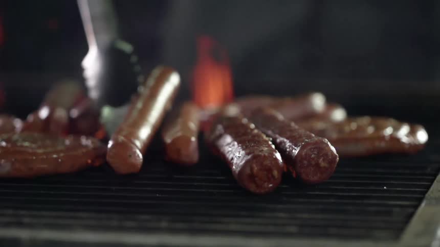 Tongs flipping a hot dog on an open flame grill
