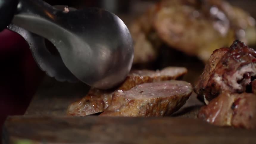 Metal tongs place a piece of cooked meat on a wooden platter