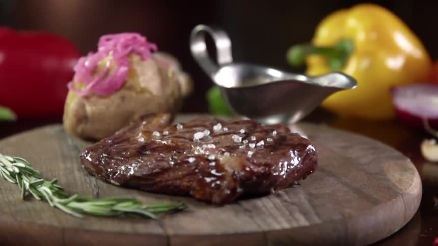 Large grains of salt falling onto a cooked steak next to a baked potato and bowl of gravy