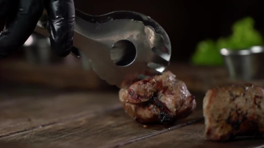 Grilled rolls of meat placed on a wooden table with metal tongs