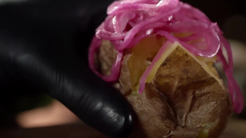A gloved hand places a baked potato with onion string topping on a board