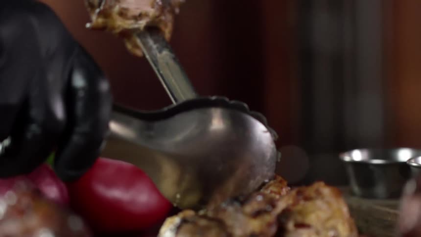 Hands with black gloves remove shish kebab from a metal rod with a pair of bigtongs