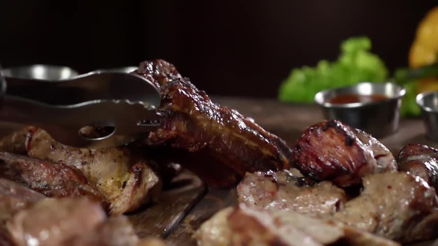 Grilled meat served with tongs on a wood table near sauces and vegetables