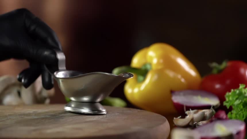 A gloved hand places a silver gravy bowl on a round wooden board lying surrounded by vegetables on a table