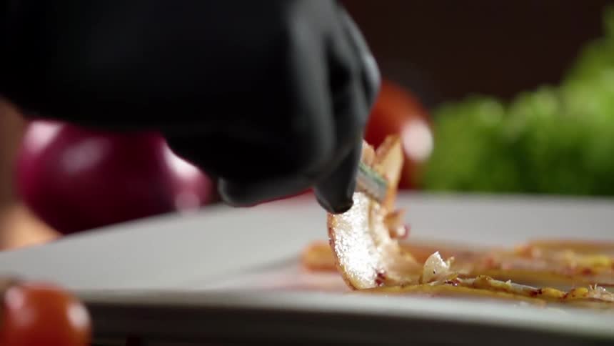 A gloved hand places a thinly sliced meat onto a plate with tongs