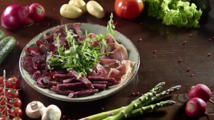 Vegetables fall atop meat in a white bowl on a table where it is surrounded by whole vegetables