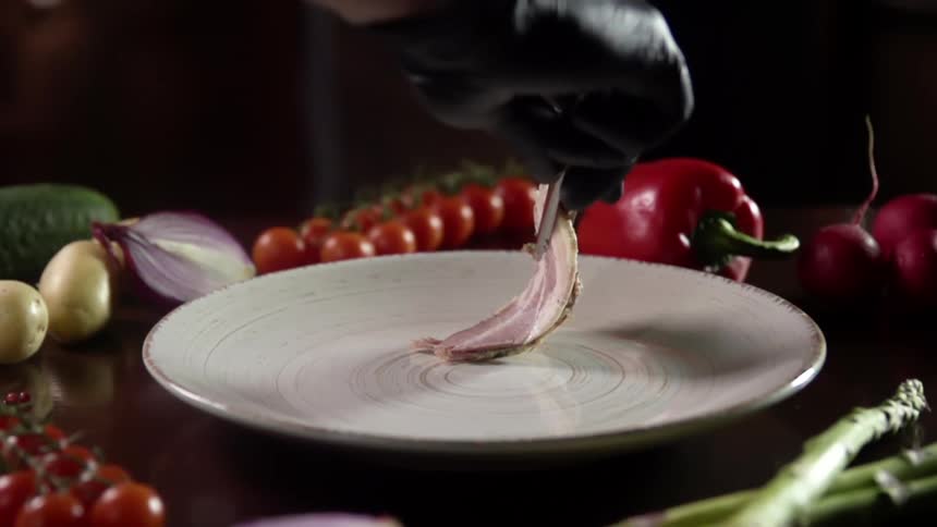 A person wearing a black glove drops a piece of thin meat on a white plate with tongs