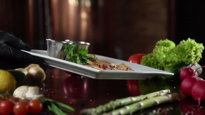 A gloved hand places a plate on to a table with vegetables surrounding the area