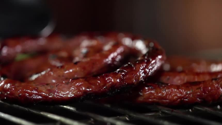 A long spiraling link of sausage being placed on a grill with a pair of tongs