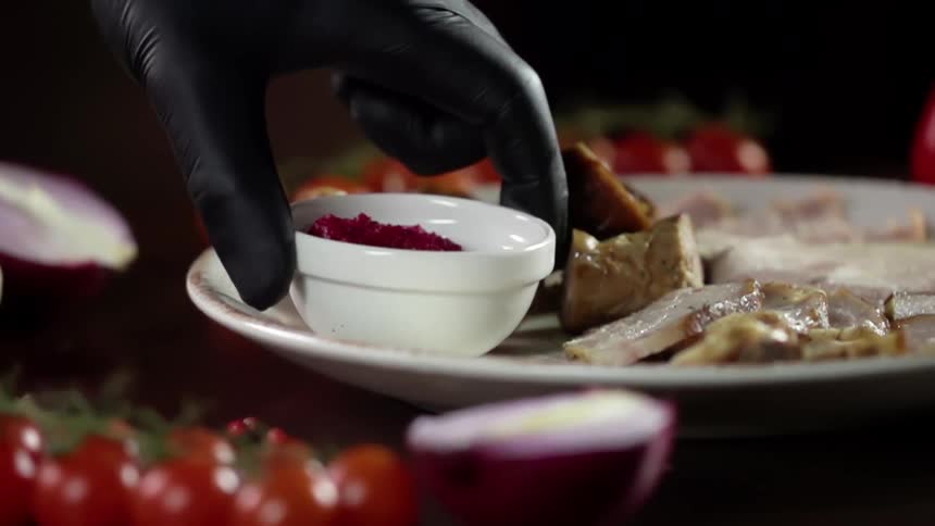 A person wearing a black disposable glove places a small bowl onto a plate with slices of cooked meat already on it