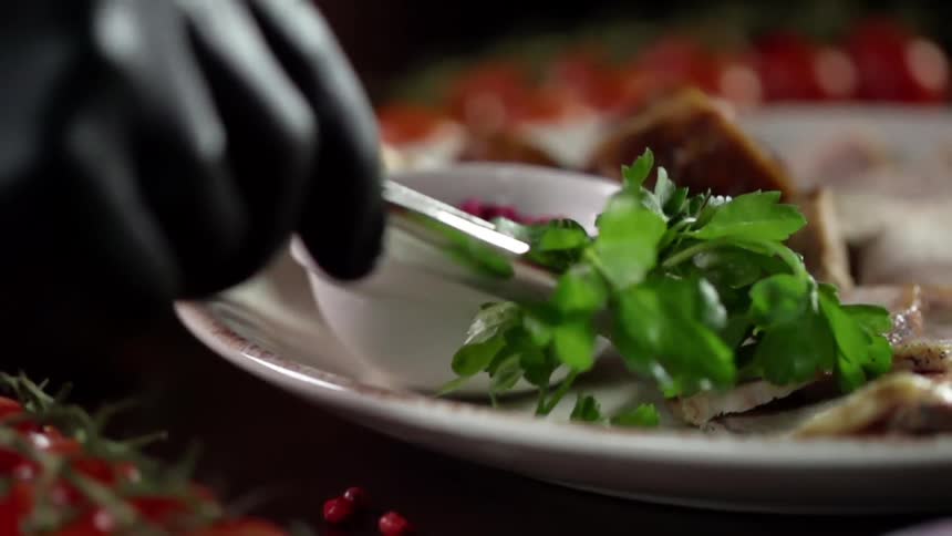 A person with gloves using a utensil to place basal on a plate with pork on a table