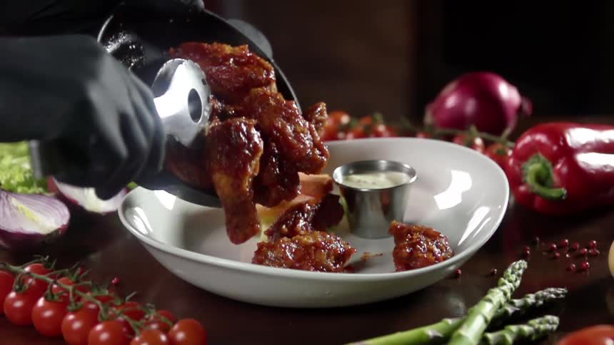 A gloved hand slides buffalo wings from a pan and onto a white plate