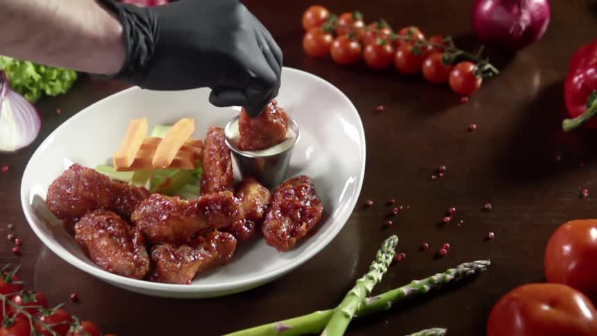 A white man wearing a black glove dips a piece of glazed meat into a dipping sauce