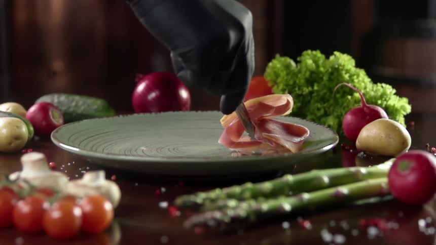 Chef wearing black latex gloves places a slice of cured meat in a ceramic plate